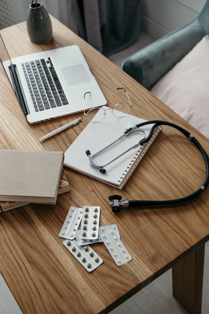 Medical stethoscope and pills on a wooden desk beside a laptop, books, and notepad.